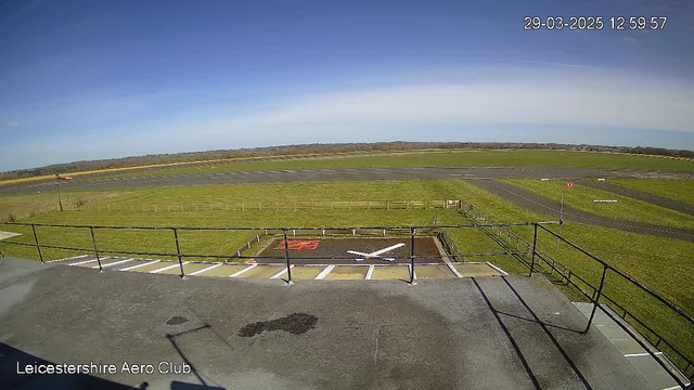 A clear blue sky stretches overhead with minimal clouds. Below, a wide runway is visible, bordered by freshly cut green grass and a fence. In the foreground, there is a helicopter landing pad marked with a large white cross. There are no aircraft visible on the runway. The image is taken from an elevated position at the Leicestershire Aero Club.