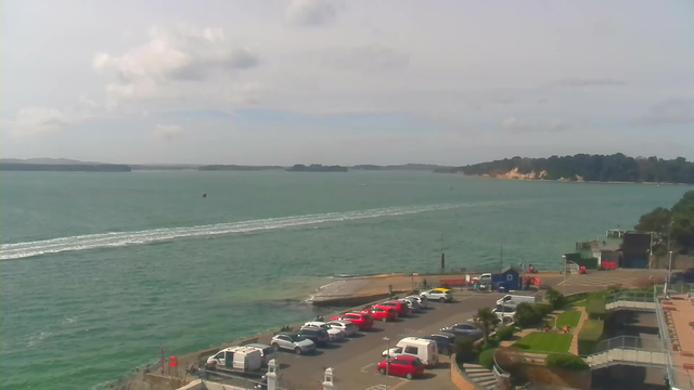 A scenic coastal view featuring a wide expanse of turquoise water with gentle waves. In the foreground, there is a parking area with various parked vehicles, predominantly white and red, along a concrete dock. To the right, a small area of greenery with landscaped plants is visible, leading to the shoreline. In the distance, green hills and a sandy cliff can be seen under a partly cloudy sky.