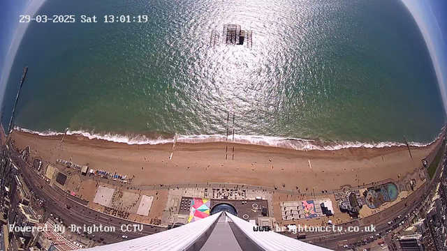 A high-angle view of a sandy beach along a coastline. The beach features a few scattered people, colorful umbrellas, and beach huts. An amusement park is visible on the shore with rides and attractions, while a pier juts out into the sea at the center of the image. The ocean appears calm, reflecting sunlight, and there are gentle waves lapping at the shore. The sky is clear with a few wispy clouds. The timestamp on the image indicates it was taken on March 29, 2025, at 1:01 PM.