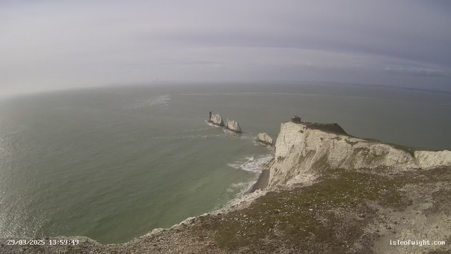 A panoramic view of a coastal landscape featuring white chalk cliffs and the sea. In the foreground, the cliffs are rugged with sparse green vegetation. Toward the center, there are several prominent sea stacks protruding from the water, with one stack having a small structure on top. The sea appears calm with gentle waves, and a light overcast sky provides a soft illumination to the scene.