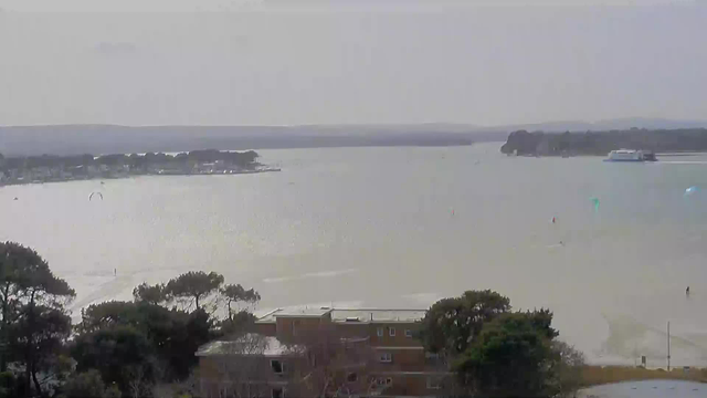 A calm body of water stretches across the view, with a gentle shimmer reflecting sunlight. In the background, hills can be seen fading into the distance. Various boats are scattered on the water's surface, and several islands are visible near the shore. Trees line the lower part of the image, providing a natural frame. The overall atmosphere is tranquil and scenic.