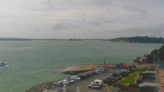 A view of the water with gentle waves and a cloudy sky. In the foreground, there is a parking area filled with various cars. A wooden dock extends into the water, and several people are visible nearby. In the background, there are hills and a coastal line with trees.
