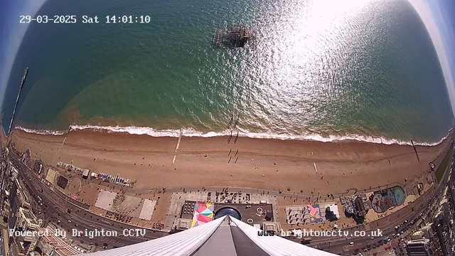 Aerial view of a beach scene taken from a webcam. The image shows a sandy beach with a curved shoreline, where gentle waves lap at the shore. Several people can be seen walking along the beach, and some are scattered on the sand. There are colorful beach umbrellas and sunbathing areas. In the water, a structure extends into the sea, possibly a pier or platform. The sun reflects brightly on the water's surface, creating a shimmering effect. The scene is set on a clear day with blue sky above.