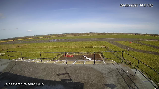 A clear view of an airport runway under a blue sky with some clouds. In the foreground, there is a flat rooftop with a railing. Below, a grassy area bordered by wooden fences leads to a runway where a small airplane is visible taxiing. The background features distant hills and scattered clouds. A sign indicating the Leicestershire Aero Club is present at the bottom left.
