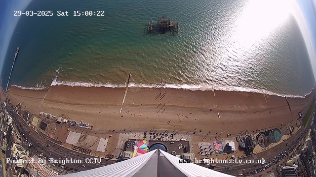 An aerial view shows a sandy beach with a bustling shoreline and gentle waves. People walk along the beach, while colorful beach huts and structures are visible below. A pier extends into the water on the left side, with a sunlit ocean reflecting light. The scene captures a lively atmosphere typical of a seaside location.