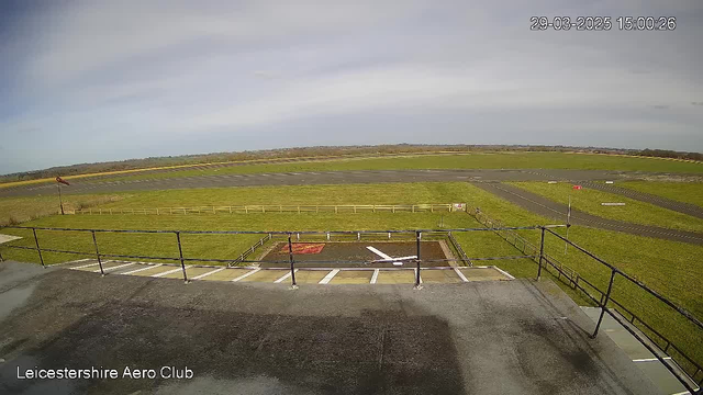 A wide view of an aerodrome with a grassy runway and a wooden fence in the background. The sky is mostly clear with a few clouds. In the foreground, there is a partially visible railing and a marked area on the ground, possibly for aircraft activity. The runway is dark and runs horizontally across the landscape.