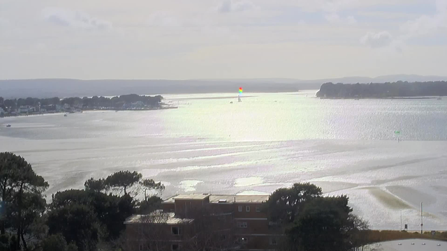 A wide view of a shimmering waterway under a cloudy sky, reflecting soft sunlight. In the distance, a small boat is visible on the water. A larger sailboat with a colorful sail, featuring red and green stripes, is sailing further out. Along the shore, trees and buildings line the waterfront, while several green and white buoys mark the water's surface. The landscape includes gentle hills in the background.