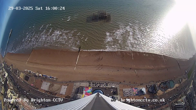 A high-angle view of Brighton Beach showing a sandy shoreline meeting the ocean. The waves create a rippling surface on the water. A pier extends into the sea, while various people stroll along the beach. Beneath the view, there are colorful beach huts and amusement park attractions visible along the promenade. The image is taken on a clear day with minimal clouds, and a timestamp indicates it is Saturday, March 29, 2025, at 4:00 PM.