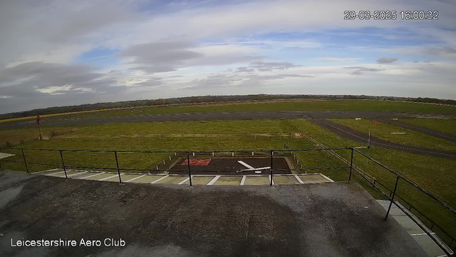 A view from a webcam overlooking a grassy field and an airstrip. In the foreground, there is a flat rooftop area with a railing, and a rectangular patch of dirt featuring a white arrow and a reddish square. The background shows a wide expanse of green grass, intersected by a paved runway. The sky is mostly cloudy with patches of blue visible. The date and time displayed in the corner reads March 29, 2025, at 16:00:32.