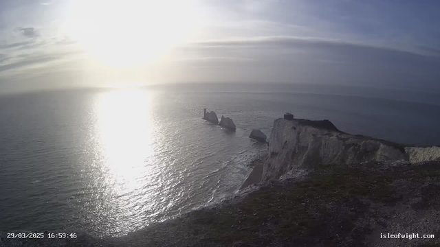 A panoramic view of the coastline at sunset, featuring a rocky shoreline extending into the calm sea. The sun is low on the horizon, casting a shimmering reflection on the water. In the distance, several large rock formations emerge from the sea, and a high cliff on the right side rises above the shoreline, partially covered in grass. The sky is a mix of soft blues and yellows, indicating the time is late afternoon.