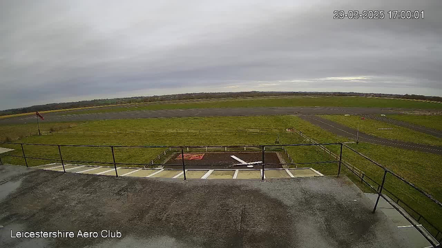 A view from a webcam overlooking an airport. The image shows a grass runway with markings and a few aircraft stands. In the foreground, there is a structure with a white object resembling a plane wing on the ground. A small red building is visible near the edge of the runway. The sky is overcast with gray clouds, and there is a light breeze indicated by a flag on a pole. The scene gives a sense of an aviation setting during daytime.