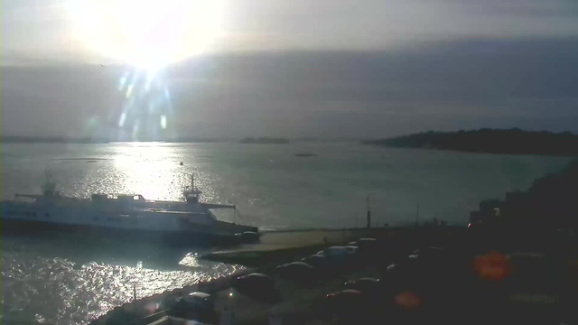 A bright sun shines in the sky, reflecting off the water of a calm harbor. A white ferry is docked at a pier on the left side of the image. The ocean appears shimmering and silver as the light bounces off its surface. In the foreground, several cars are parked along the shoreline, with a faint outline of land visible in the distance on the right side. The sky is mostly cloudy, giving a soft, muted atmosphere to the scene.