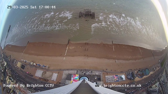 A high-angle view of a beach and coastline with the sea in the background. The sandy beach stretches horizontally across the image, dotted with small figures, likely people. In the sea, gentle waves are visible. A dilapidated pier emerges from the water, and several wooden posts are aligned parallel to the shore. Below, various structures including businesses and attractions can be seen, with some colorful patterns on the ground. The image captures a cloudy sky above and appears to be taken from a tall structure. The time and date are displayed in the top corner.
