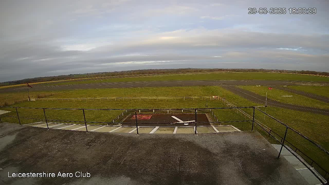 A view from a webcam at Leicestershire Aero Club shows a grassy airfield under a cloudy sky. In the foreground, there is a helipad with a white cross marking the center, surrounded by a wooden fence. The background features runways and grassy areas with distant hills visible on the horizon. The left side of the image includes a wind sock indicating wind direction, and the time and date are displayed at the top right corner.