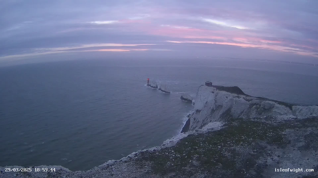 A coastal scene at dusk featuring a rocky shoreline with white cliffs. In the distance, a red lighthouse stands along the water's edge, guiding passing ships. The sea appears calm, with gentle waves reflecting the soft pastel colors of the evening sky, which transitions from light blue to pink and gray. A small structure can be seen near the lighthouse, perched on the cliff. The date and time are displayed in the bottom left corner, indicating the scene was captured on March 29, 2025, at 6:59 PM.