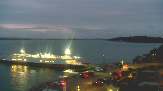 A ferry is docked at a harbor during twilight. The ferry is illuminated with bright lights, reflecting on the water. In the foreground, several parked cars are visible, with some headlights on. The background shows a calm body of water and distant land on the horizon under a cloudy sky.
