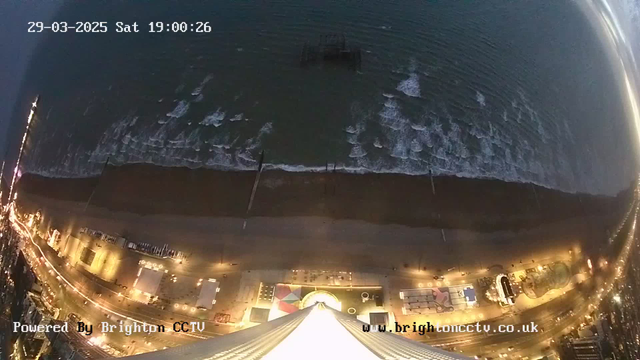 Aerial view of a coastline at dusk, featuring a sandy beach and rolling waves. The water meets the shore, creating white foam where the waves crash. An old pier is visible in the water, partially obscured. Bright lights line the beach, illuminating various structures and attractions. A colorful carousel shape is noticeable near the bottom of the image. The sky is darkening with some distant lights from the beachside. The date and time is displayed at the top of the image.