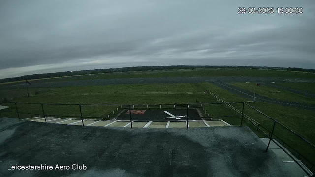 A cloudy sky is visible above a large grassy field with a runway. In the foreground, there is a fenced area with what appears to be a helipad marked by a red square and a white arrow. The ground is mostly gray, likely a structure or path, with a railing visible along the edge. The scene is serene and open, with no visible aircraft or vehicles.