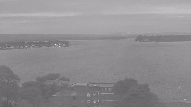 A view of a calm body of water, bordered by land on both sides. In the foreground, there are trees and a building with several windows. The water stretches into the distance, where land and hills are visible under a cloudy sky. The scene appears monochrome, conveying a tranquil and overcast atmosphere.