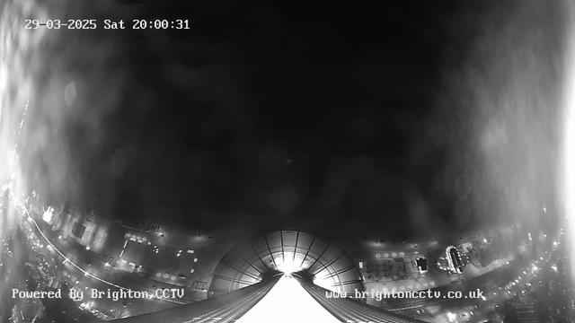 A nighttime view captured from a webcam shows a cityscape illuminated by streetlights and lights from buildings. The image is primarily black and white, with a noticeable upward angle, highlighting a structure with a round dome at the top. The bottom portion of the image features a roadway and various building outlines, with the date and time displayed in the top left corner. The scene appears somewhat foggy or hazy, slightly obscuring details.