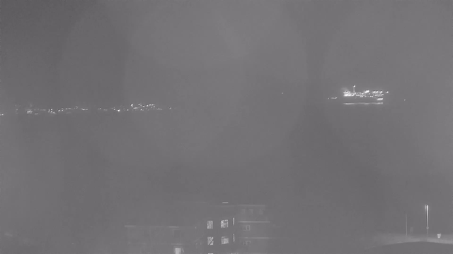 A dimly lit night scene shows a body of water with faint lights on the horizon, suggesting a distant shoreline or city. In the foreground, there is a dark building with some illuminated windows. A larger illuminated object, possibly a boat or ferry, is visible on the water, slightly to the right. The overall atmosphere is dark and somewhat foggy, with soft circular blur effects in the image.