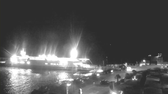 A brightly lit ferry is docked at a pier, reflecting light on the water's surface. The scene is set at night, with minimal background light beyond the ferry. Various vehicles are parked near the edge of the pier, and there are some palm trees visible in the foreground.