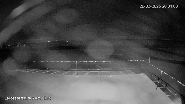 A nighttime view from a webcam at Leicestershire Aero Club. The image is predominantly dark with a faint outline of a fence along the edge of a flat surface, possibly a runway or a viewing platform. In the distance, very faint lights are visible, suggesting the presence of buildings or streets. The scene appears cloudy and slightly blurred, possibly due to water droplets on the lens. The date and time displayed in the top right corner indicate March 29, 2025, at 20:01.