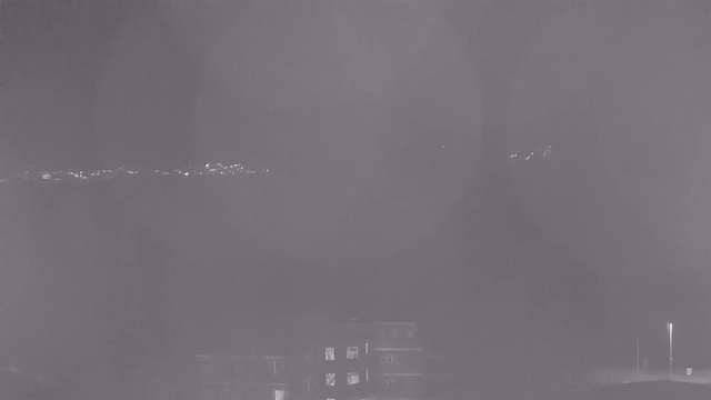 A monochromatic webcam image showing a dark landscape with faint outlines of buildings in the foreground. The background features a horizon with soft, dim lights which may represent distant structures or reflections. Overall, the scene is hazy and lacks clarity, creating a moody, nighttime atmosphere.
