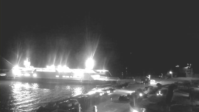 A brightly lit ferry is docked at a harbor at night, with strong lights shining from its top and side. The water reflects the lights, creating a shimmering effect. In the foreground, several parked cars line the shore, and faint outlines of structures and other vehicles are visible in the dark background. The scene conveys a peaceful yet active ambiance of a waterfront at night.
