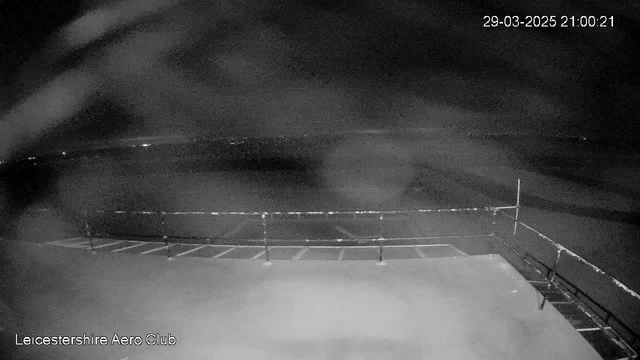 The image shows a dark scene from a webcam at the Leicestershire Aero Club. In the foreground, there is a flat surface, possibly a parking area, with faint outlines of parking spaces. A low railing runs across the bottom of the image. In the background, there are indistinct distant lights. The overall atmosphere is quiet and nighttime with limited visibility due to darkness. The timestamp on the image reads 29th March 2025 at 21:00:21.