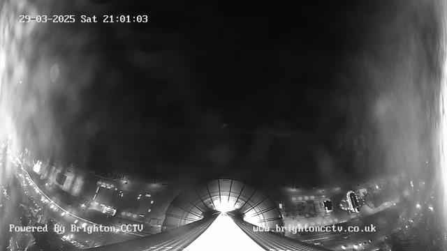 Black and white image from a webcam showing a nighttime cityscape. At the top, there is a timestamp indicating the date and time as 29-03-2025, 21:01:03. The bottom half of the image features a distant view of buildings and streets illuminated by lights, with a prominent structure appearing to lead toward the center of the image, possibly a dome or roof. The city below is partially obscured by a mist or smoke effect, creating a cloudy atmosphere. The watermark at the bottom shows "Powered By Brighton CCTV" and the website "www.brightoncctv.co.uk."