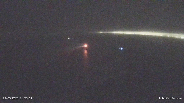 A dark scene captured from a webcam shows a body of water at night with limited visibility. In the foreground, a small boat emits a faint orange light, while another boat with a blue light is visible nearby. The horizon is lined with a distant strip of light indicating the presence of land, partially obscured by darkness. The timestamp at the bottom reads 29/03/2025, 21:59:51.