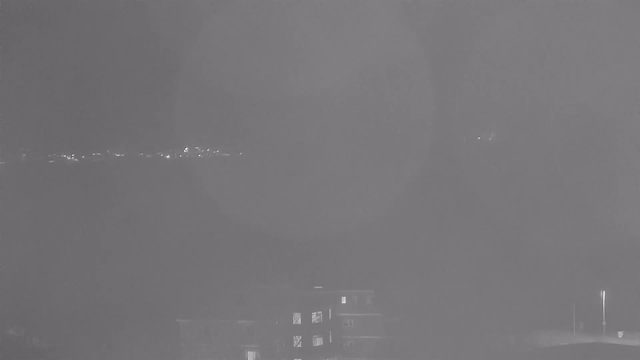 A dimly lit urban scene shows an outline of a building in the foreground with several lit windows. In the background, faint lights indicate the presence of a distant cityscape or shoreline. The overall atmosphere is hazy, likely due to fog or low visibility conditions, with a soft glow emanating from the lights.