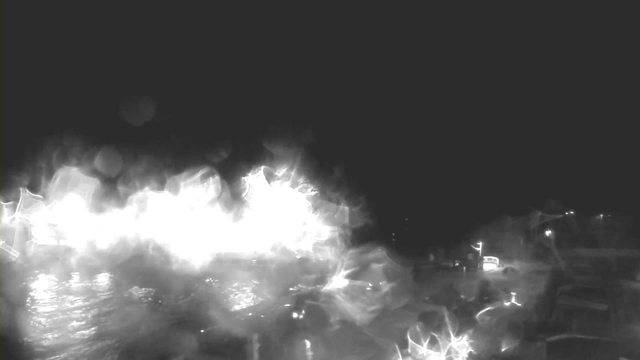 A dark scene with bright, glowing areas reflecting off a body of water. The lights appear to be scattered, suggesting possible structures or objects along the shoreline. The overall atmosphere is hazy and somewhat mysterious, with indistinct shapes in the foreground.