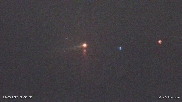 A dark nighttime scene showing faint lights reflecting on water. On the left, a small white light is visible, possibly a distant star or boat light. Toward the center, a larger orange light stands out, possibly from a nearby vessel or buoy. To the right, there are two small blue lights, also reflecting on the water. The overall atmosphere is low visibility, hinting at a quiet marine setting. The image includes a timestamp at the bottom left indicating the date and time.