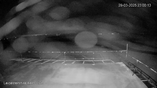 A dark scene captured from a webcam at the Leicester Aero Club, showing a flat surface with a railing in the foreground. The background appears dim with scattered lights indicating distant objects or structures. The time and date are displayed in the upper right corner: 29th March 2025 at 23:00:13.