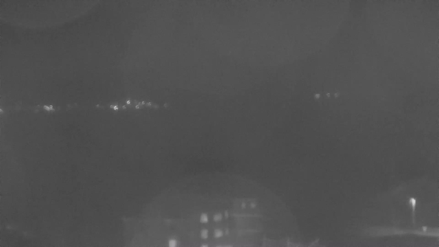 A dimly lit scene featuring a body of water with faint lights reflecting off its surface in the distance. The foreground shows an indistinct outline of a building, while the background is a dark expanse, possibly indicating the presence of hills or trees. The overall atmosphere is quiet and nighttime, with limited visibility.