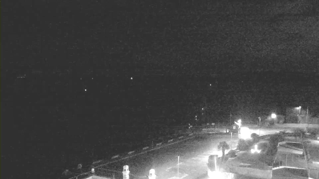The image shows a dark scene with faint lights in the distance, possibly reflecting off water. In the foreground, there are outlines of structures, including what appears to be boats or vehicles, and some illuminated objects indicating artificial lighting. The sky is mostly black with minimal visibility, suggesting nighttime.