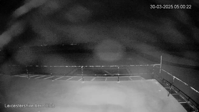 A dark scene is captured from a webcam at the Leicestershire Aero Club. In the foreground, there is a flat surface, likely a tarmac area, with markings indicating parking spaces. A low railing runs along the edge, with a vertical post on the right. The background is mostly empty, with a faint glow of lights in the distance, suggesting an urban area or some distant objects. The overall image is dimly lit, emphasizing the lack of visibility.