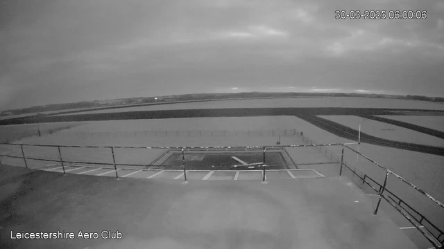 A black and white image from a webcam showing a wide view of a flat area with a faint horizon line in the distance. In the foreground, there is a helipad outlined on the ground, marked with a white "H" symbol. There are horizontal lines indicating potential runway markings running through the scene. The sky is overcast with clouds, creating a dim ambiance. The date and time in the top right corner reads March 30, 2025, at 6:00 AM. The lower left corner displays the text "Leicestershire Aero Club."