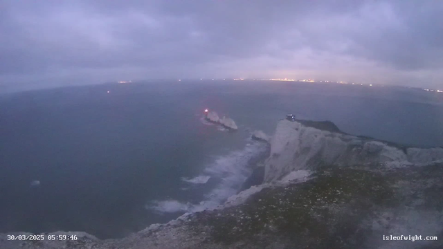 A cloudy sky hangs over the ocean, with dark waters and gentle waves lapping against a rocky shore. In the foreground, a cliff is visible, covered with sparse greenery and stones. To the left, there is a lighthouse on a small rock formation, emitting a red light. In the distance, faint lights from a shoreline city can be seen along the horizon, indicating human activity. The overall mood is muted, with a diffuse light suggesting either dawn or dusk.