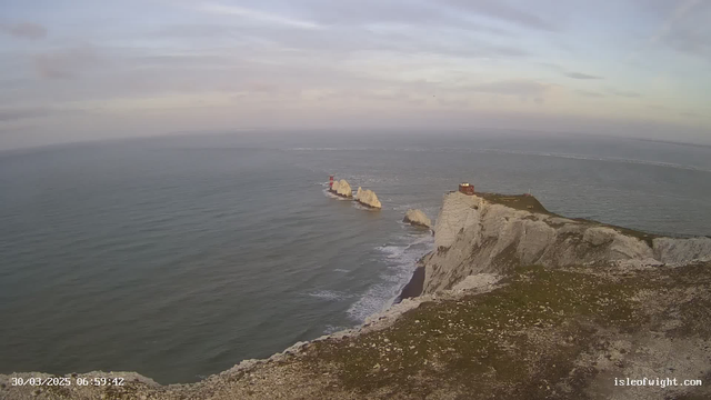 A panoramic view of a coastline during dawn, featuring rocky cliffs and the ocean. To the left, there are three prominent white rocky outcrops in the water with a red lighthouse on the first one. The sea is calm with gentle waves lapping against the shore. In the upper right corner, there is a small red structure atop the cliffs. The sky is cloudy with soft pastel tones, and the scene conveys a serene atmosphere.