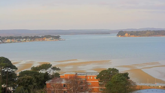 A serene landscape featuring a calm body of water, with gentle waves reflecting the light. In the foreground, there are scattered trees and a building with large windows. Across the water, a small coastal town with various structures and a noticeable marina can be seen, along with boats docked. In the distance, a small island with a castle-like structure is visible against a backdrop of rolling hills and a pale sky. The scene conveys a peaceful, natural environment.