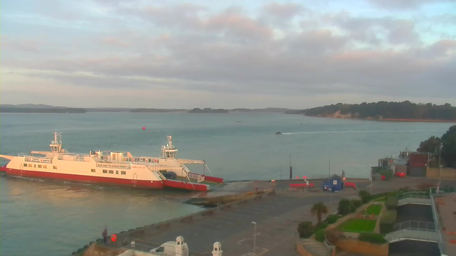A view of a harbor during the early morning. A large white and red ferry is docked near the shore, with the water gently rippling around it. In the background, there are green hills and a coastline with trees. A small motorboat is seen moving across the water. The sky is partly cloudy with soft pastel colors, indicating a calm atmosphere. There are pathways, some planters, and structures along the harbor's edge.
