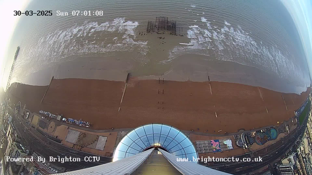 A high-angle view from a webcam looking straight down towards a beach along the coast. The image shows a sandy beach with gentle waves lapping at the shore. In the center, there is a partially submerged pier structure with people walking nearby. On the left side, there are various beach amenities and attractions, with spots of colorful designs. The image is taken during early morning hours, and there is a soft light illuminating the scene. The date and time are displayed at the top left corner, indicating March 30, 2025, at 07:01:08.