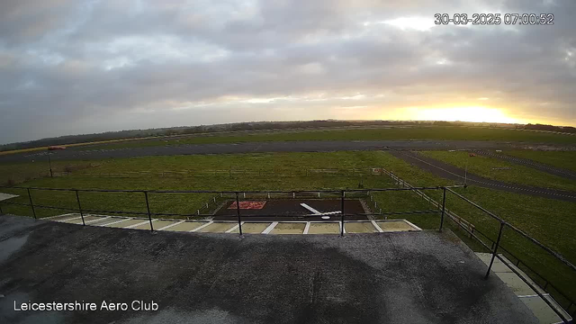 A grassy runway stretches out under a cloudy sky, with a hint of sunrise visible on the horizon. A fenced area and a few objects are positioned near the base of a viewing platform. The scene captures a tranquil aviation environment at the Leicestershire Aero Club, with a mix of grass and gravel surfaces leading to the runway. The overall atmosphere is early morning, suggesting a new day beginning.