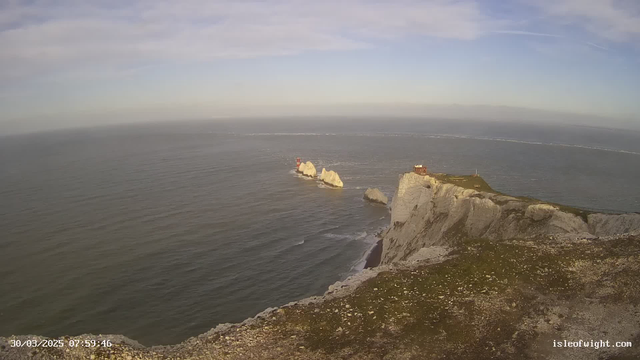 A panoramic view of a coastal landscape at dawn. In the foreground, a rocky cliff with sparse vegetation extends across the bottom of the image. Below the cliff, gentle waves roll in from a calm sea. Off to the right, a small group of white and gray rocks protrudes from the water, two of which have small red structures on top, possibly lighthouses. The sky above showcases a mix of soft blue and wispy clouds, indicating clear weather. The image is marked with a timestamp in the lower left corner.