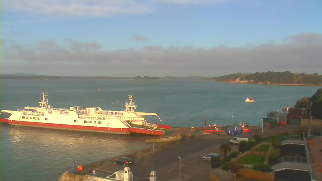 A large white ferry with red accents is docked at a port. The ferry is labeled with the words “GIVE WAY TO CHAIN FERRY.” In the background, there is a calm blue sea and distant land, with green hills. A small boat is traveling across the water, and several vehicles are parked near the port area. The sky is mostly clear, with some clouds.