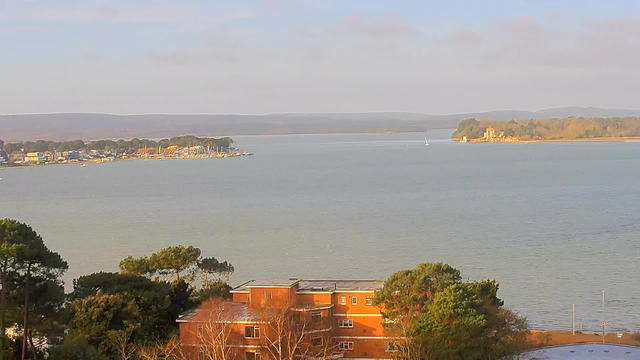 A scenic view of a calm body of water reflecting a clear sky with light clouds. In the foreground, there are trees and a building with reddish-brown walls. To the left, a shoreline features small boats and several colorful buildings. In the distance, a small island is visible, topped with greenery and what appears to be a castle or structure. The background consists of gentle hills.