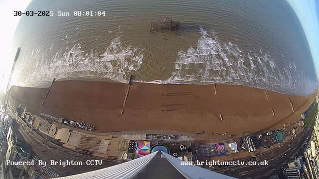 Aerial view of a beach at sunrise, with waves gently lapping against the shore. The sandy beach is visible, along with a wooden pier extending into the water. Various structures, including a carousel and other buildings, are situated near the beach. The sky is mostly clear, and the ocean appears calm with light ripples. In the foreground, a part of a building or structure can be seen, indicating the viewpoint is from a high elevation. The date and time are displayed at the top.